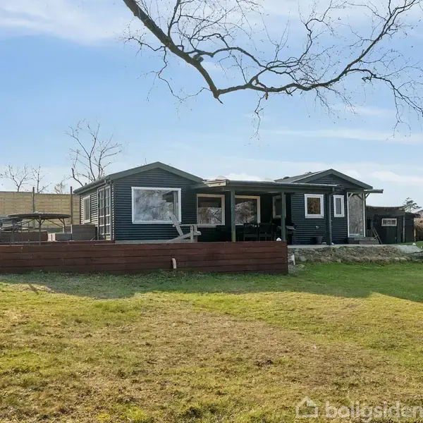 Et fritidshus står stille omgivet af græsplæne og træer, med en stor terrasse foran. Husets mørke facade står i kontrast til den klare blå himmel.