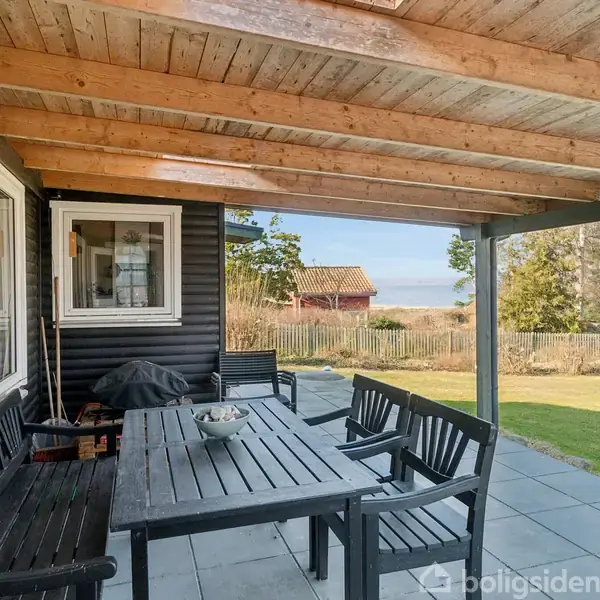 Terrassemøbler i sort træ står på en overdækket veranda. Der er udsigt til en grøn have og træbygninger bag et trægærde under en klar himmel.