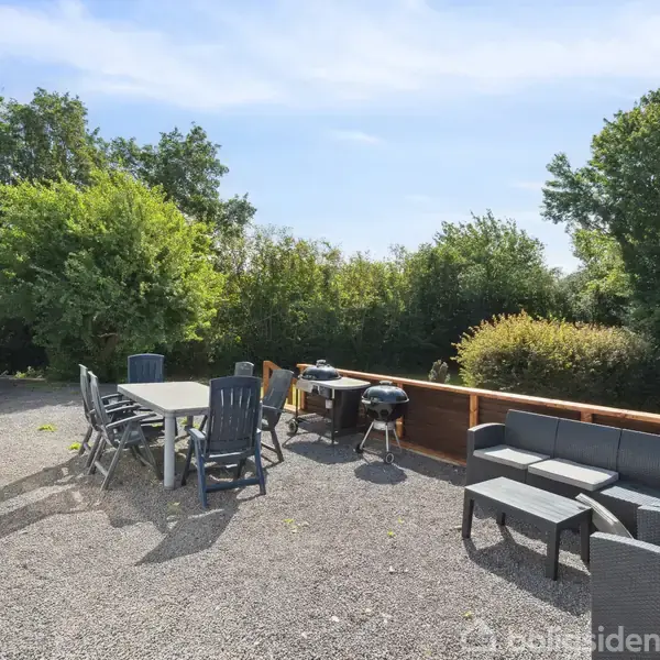 En terrasse med havemøbler og en grill står udendørs. Bord og stole er placeret ved siden af huset. Bænkesæt er ved et hegn. Omgivet af frodig vegetation.