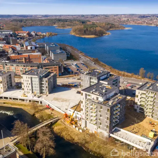 Flere moderne lejlighedsbyggerier står strategisk placeret under opførelse ved en vandkant, omgivet af en by og en stor sø med grønne øer i baggrunden.