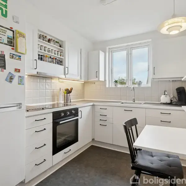 Køkken med hvide skabe og skuffer, komfur og vask på en L-formet bordplade, køleskab med magneter til venstre, og sort stol ved spisebord, i et lyst rum med vindue.