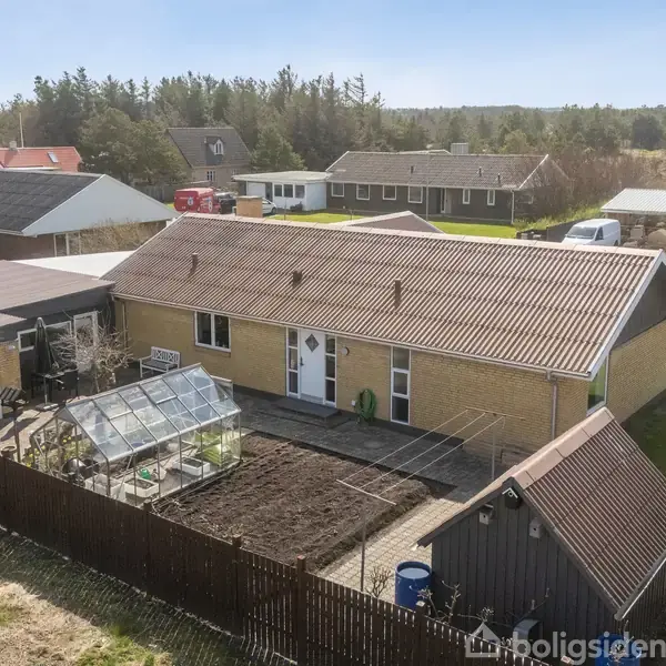 Et enfamiliehus med brun facade og rødt tag ligger i et villakvarter. Foran er der en have med et drivhus og bed. Området er roligt med flere huse i nærheden.