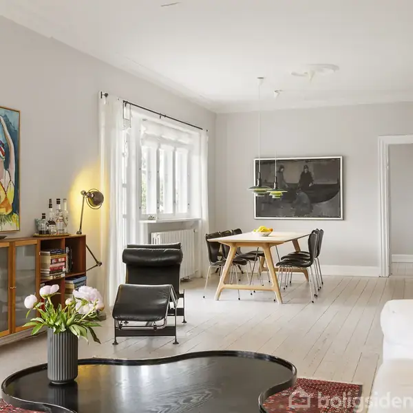 Stue med sofa, sort lænestol og sofabord; spisebord med stole i baggrunden. Væggene prydes af farverige malerier. Store vinduer lukker lys ind i det lyse, stilfulde rum.