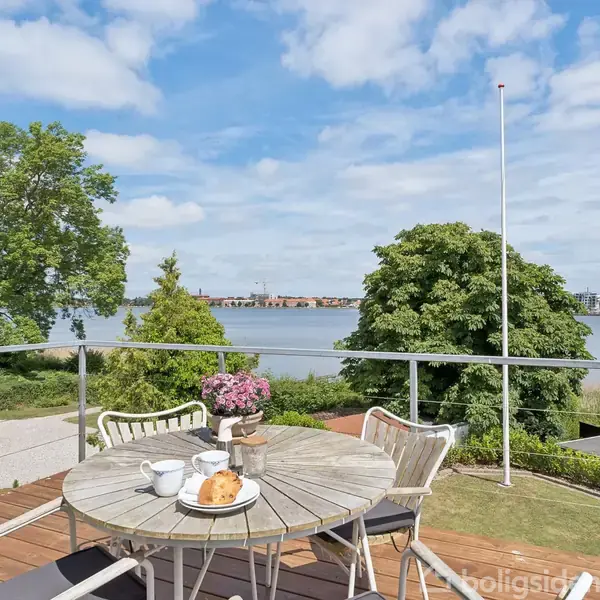 Et rundt havebord står på en træterrasse med kaffe og kage serveret. Bordet er omgivet af stole. I baggrunden ses en have, træer, en sø og en by i horisonten.