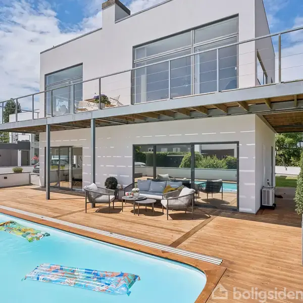 Moderne villa med to etager, en stor balkon og en terrasse med pool, omgivet af grønne planter. Poolen har en oppustelig madras, og møblerne på terrassen skaber en hyggelig atmosfære.