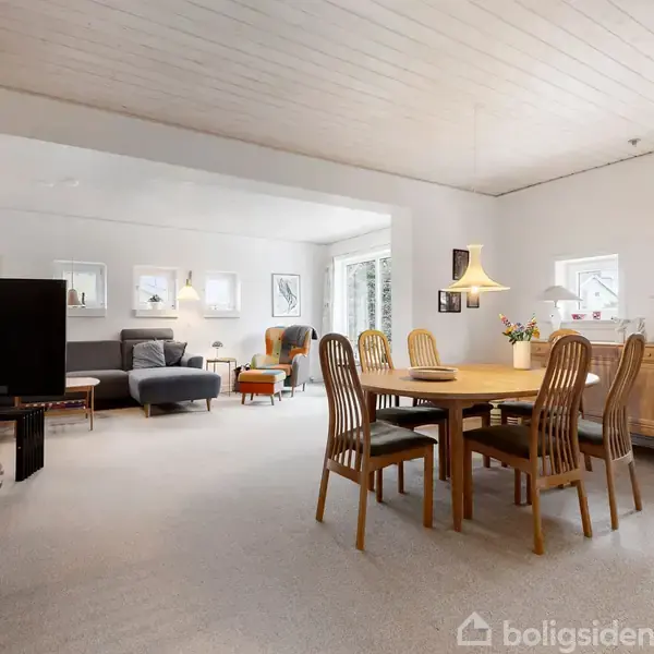 En stue med et spisebord i træ og seks stole står midt i rummet. En sofa og lænestol ses i baggrunden sammen med en TV-enhed. Hylder og skænk langs væggene.