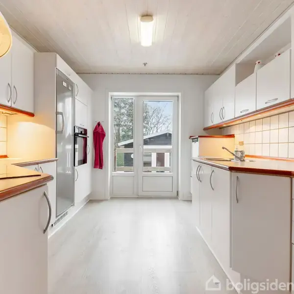 Hvidt køkken med træbordplader. Køkkenet er tomt, men oplyst, og en rød viskestykke hænger ved ovnen. Store vinduer viser en terrasse og have udenfor.