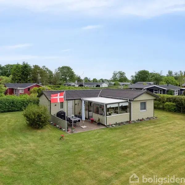 Et lille sommerhus med en dansk flag på terrassen. Det står midt i en velplejet græsplæne, omgivet af buske og træer, i et roligt sommerhusområde.