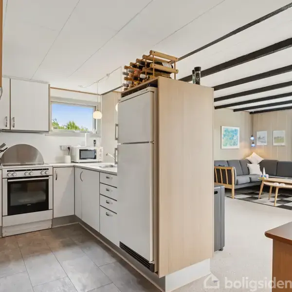 Køkken i étplanshus med komfur og køleskab. Åben forbindelse til stue med sofa og sofabord. Store vinduer giver naturligt lys. Dekorative bjælker i loftet.