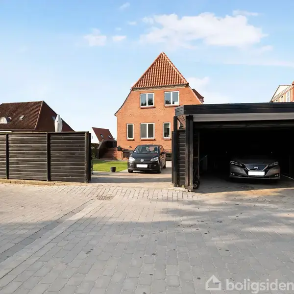 Bil parkeret i garage ved siden af åben plads. Bagved ses et rødt murstenshus med flere vinduer. Flisebelægningen strækker sig over området, omgivet af træhegn og andre huse.