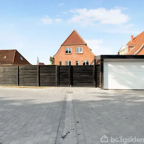 En sort trægarage med en hvid port står foran en lang træhegn. I baggrunden ses røde murstenshuse med spidse tage under en blå himmel.