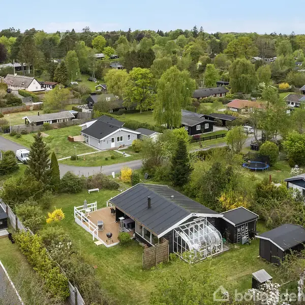 En samling af huse med haver beliggende i et frodigt, grønt boligkvarter. Et specifikt hus er fokuseret, omgivet af et drivhus og græsklædte områder. Veje er synlige imellem ejendommene.