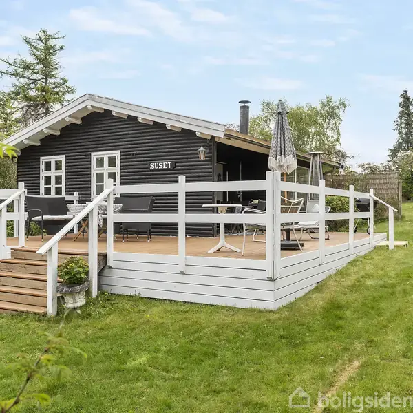 Sort træhus med skilt "SUSIE" på facade. Huset har en stor veranda med hvide gelændere, havemøbler og en parasol. Det omkranses af en grøn græsplæne og træer.