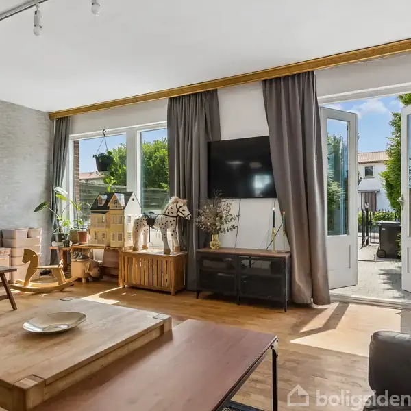 Et lyst, moderne stueområde med trægulv og stort vinduesparti. Et TV hænger på væggen, omgivet af reoler og legetøjshuse. Dørene åbner ud til en terrasse.