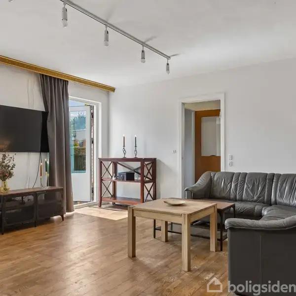 Stue med sort lædersofa og træbord, vendt mod en væg med fladskærm og dør til balkon. Store vinduer med gardiner slipper lys ind.