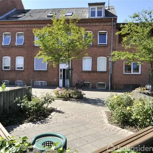 En række røde murstenshuse ses med sollys i gårdmiljø. To bænke er placeret langs flisebelagt sti, omringet af blomsterbede og træer. Cykel lænet op ad mur.