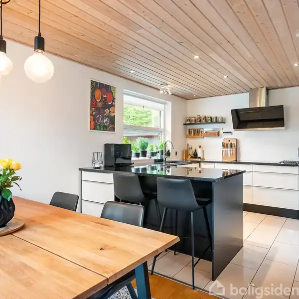 Køkkenbord omgivet af sorte stole; træloft med hængende lamper; træbord med vase med gule blomster; køkkenvask ved vindue; vægkunst, hvide skabe, emhætte i moderne køkkenindretning.