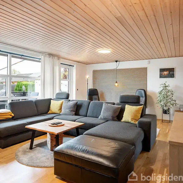 En stor, grå hjørnesofa står i en moderne stue med trægulv og træloft. Sofaen har pyntetæpper og puder. Ved siden af er et lavt bord. Rummet er lyst med store vinduer.