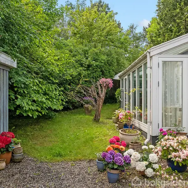 Et drivhus med glasvægge står ved siden af en grussti omgivet af farverige blomster i potter. I baggrunden er der en frodig have med grønne træer og buske.