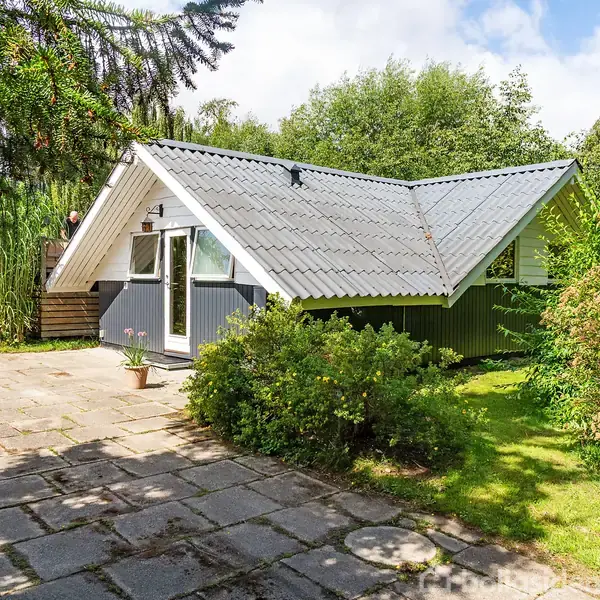 Sommerhus: Gråt og hvidt hus med skråtag ligger omgivet af grønne buske og græsplæne, set fra en flisebelagt terrasse.