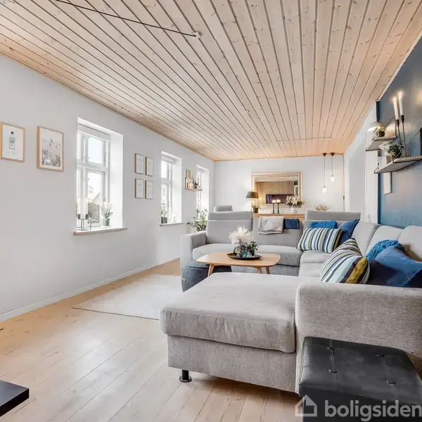 En moderne stue med en stor grå hjørnesofa fyldt med puder. Sofaen står op ad en væg med indrammede billeder. Rummet har et lyst trægulv og loft med flere vinduer på siden.