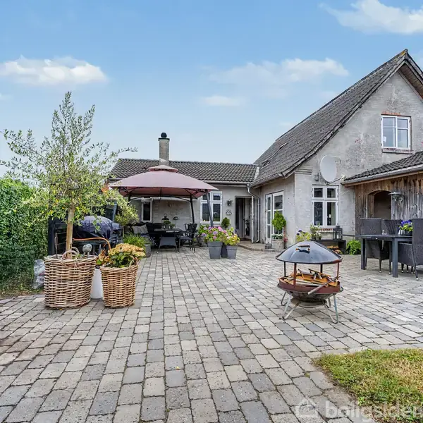 Terrasse med flisebelægning, hvor havemøbler og en parasol står i midten. Omkranset af grønne planter op ad en ældre, charmerende bygning med et murstens- og træbeklædt udhus.