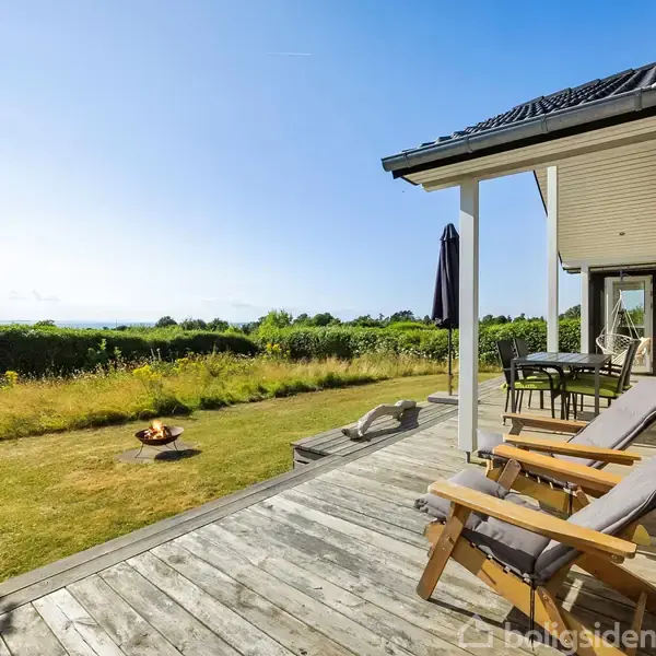 Træterrasse med to komfortable liggestole; placeret ved et hus med udsigt til grøn have og horisonten. Et udendørs spisebord står under en overdækning.