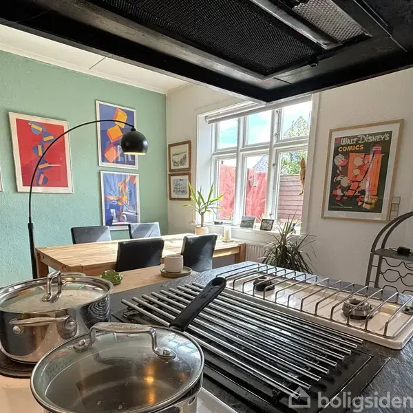 Gryder står på komfuret i et køkken/alrum. I baggrunden er der et spisebord med stole, farverige kunstværker på væggen og en stor lampe. Sollys strømmer ind gennem vinduerne.