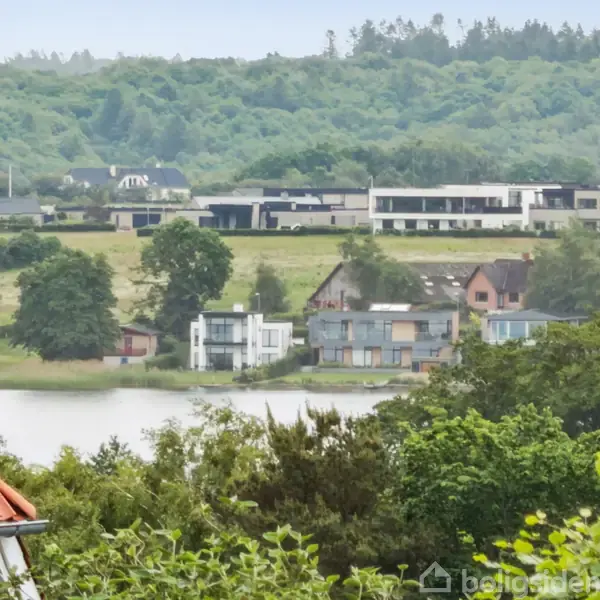 Røde hustage og moderne boliger står langs en sø, omringet af frodig vegetation og grønne bakker i baggrunden. Der ses en række bygninger i det fjerne.