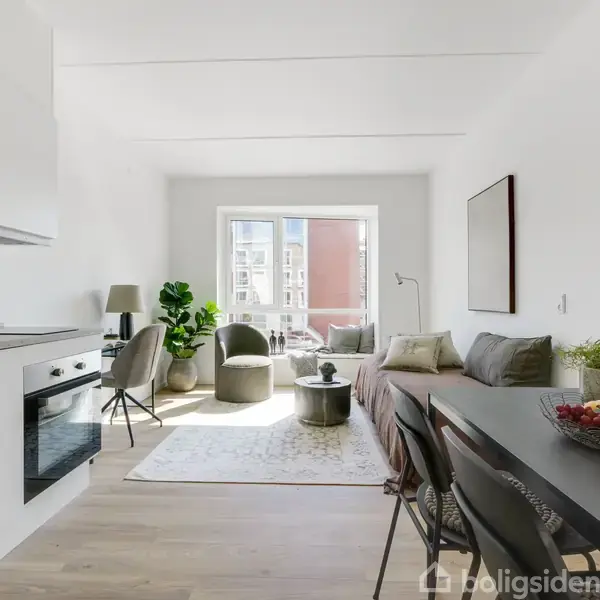 En lys stue med et moderne køkken til venstre, sofa og stole ved et vindue. En skål med frugt står på spisebordet. Rummet er stilfuldt indrettet.