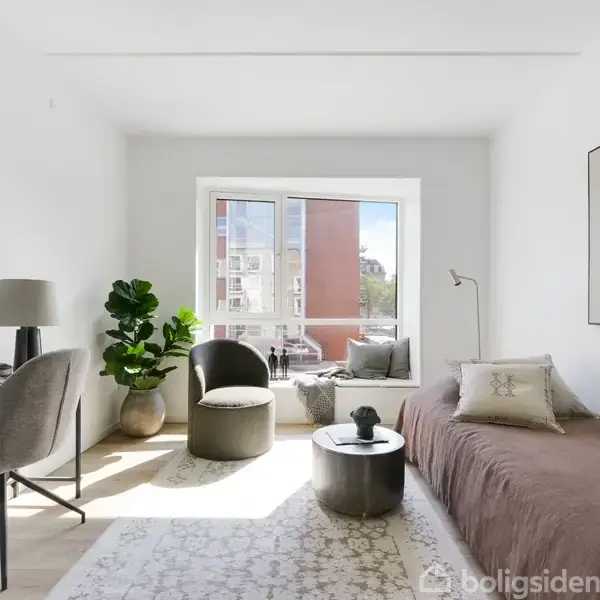 Et moderne, lyst værelse med en brun sofa, skrivebord, grå stol, potteplante og stort vindue. Et rundt bord står foran sofaen. Værelset har minimal indretning.