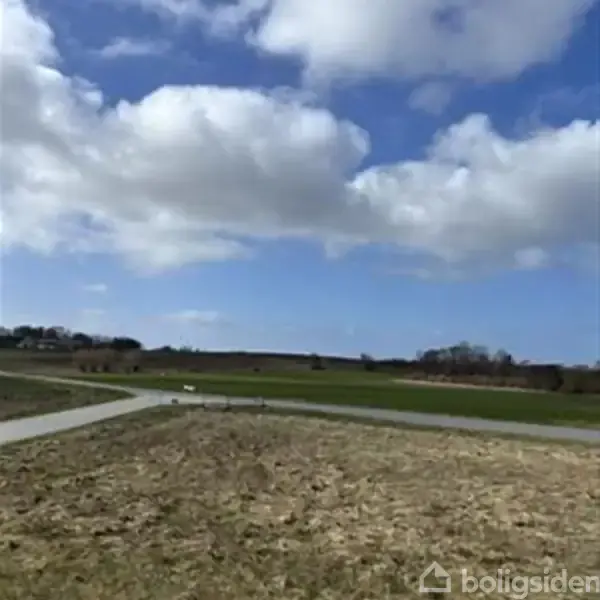En grusvej snor sig gennem et fladt, åbent landskab med grønne marker under en blå himmel med spredte skyer.