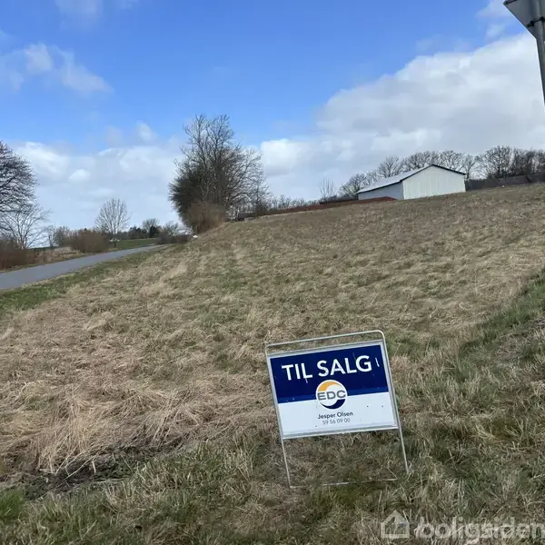 Et skilt står foran en græsmark med teksten "TIL SALG EDC Jesper Olsen Sydals." I baggrunden ses en landevej og en hvid bygning under en blå himmel med skyer.