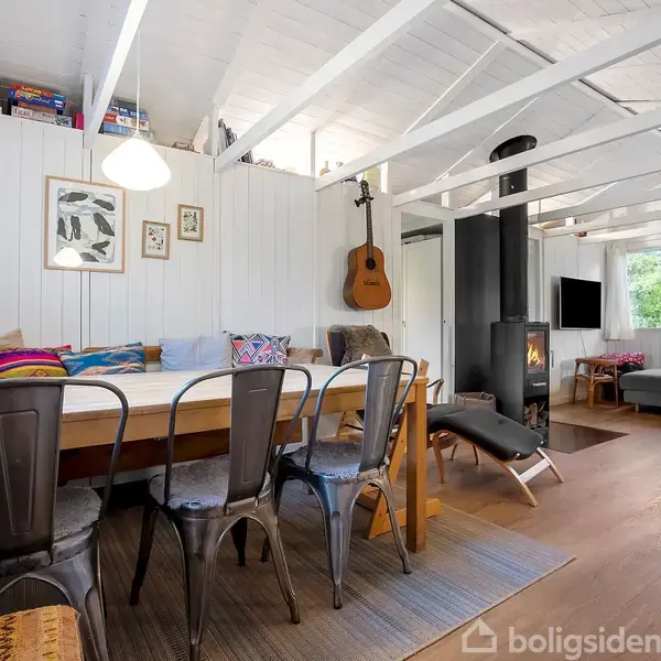 Stue med træloft og fremtrædende bjælker. Rundt spisebord med metalstole og sofa i baggrunden. Væghængt guitar og brændeovn. Store vinduer giver lys. Spil og bøger øverst på hylderne.