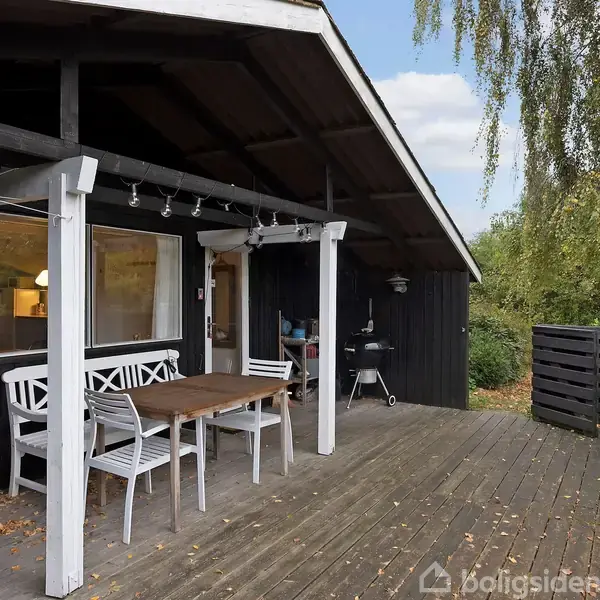 På en træterrasse står et bord og hvide stole under en sort træveranda med lyskæde. En grill ses i baggrunden. Omkring er der grønne træer.
