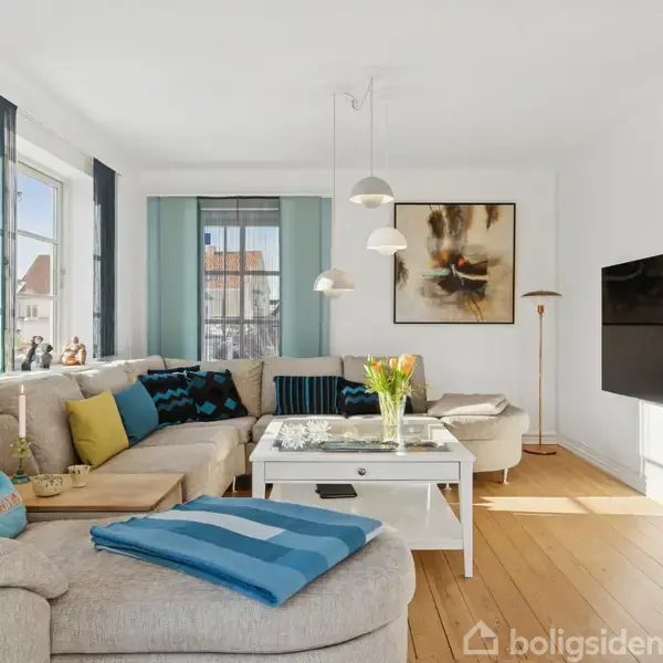 En lys stue med sofaer i beige og blå puder, lyse trælaminatgulve, et stort fladskærms-TV på væggen og et sofabord med blomster i vase. Sollys ind ad vinduerne.