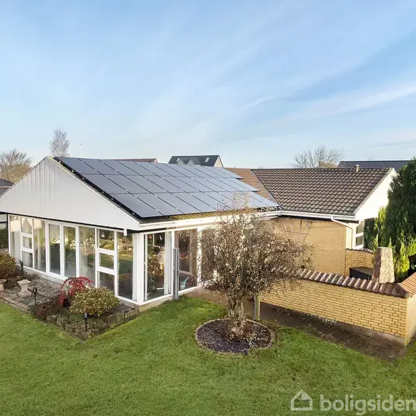 Hus med solpaneler på taget står i solskin omgivet af grøn græsplæne, buske og træer i en forstadshave.