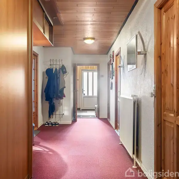 Gang med rødt tæppe fører til en dør i den fjerne ende. Vægge har knager med overtøj, og der hænger et spejl. Dørene på begge sider er af træ.