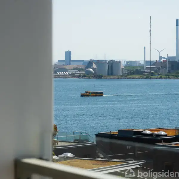 En lille båd sejler på en bred flod eller kanal. I baggrunden ses industrielle bygninger og vindmøller, mens forgrundens bygning har grønne tage.