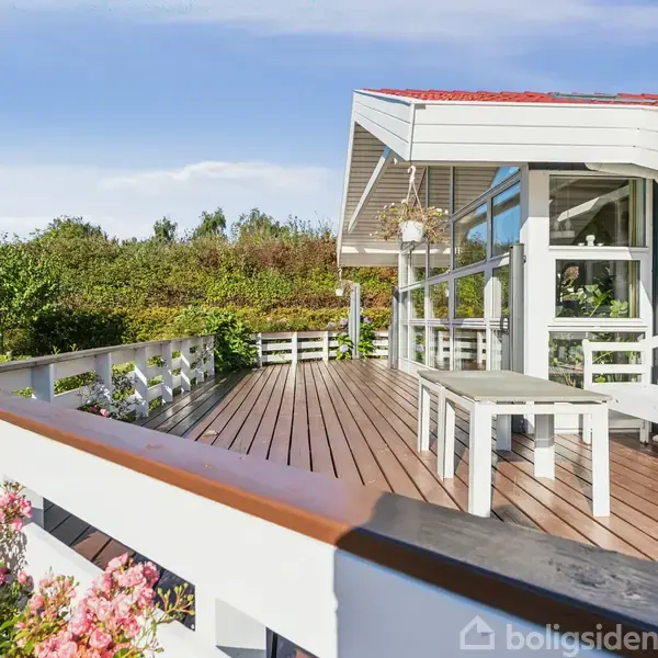 Træterrasse med hvide møbler står foran et moderne hus med store vinduespartier. Omgivet af blomster og grønne buske på en solrig dag.