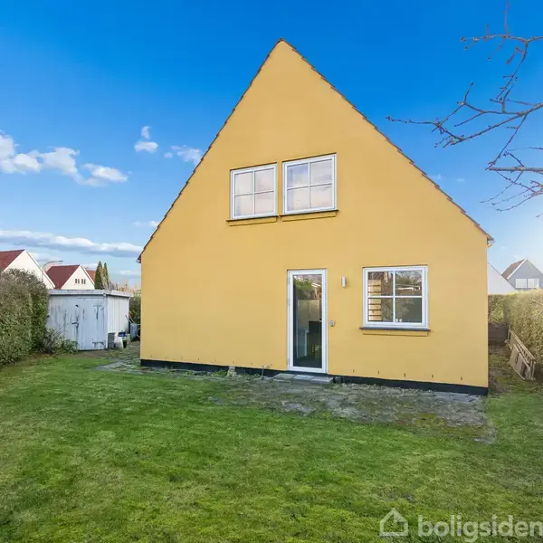 Et gult hus med to vinduer og en dør vender ud mod en grøn græsplæne. Omgivet af hække og en blå himmel. Bænk ses til venstre.