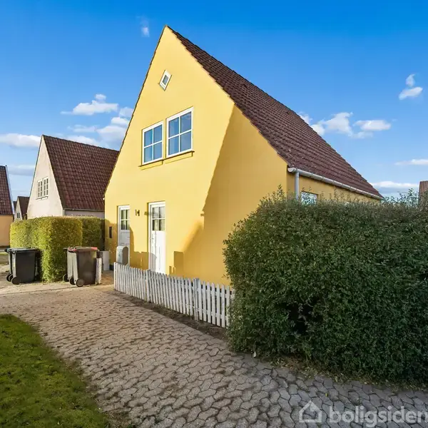 Gult hus med spidst tag står i en solrig indkørsel, omgivet af grønne hække. I forgrunden ses affaldscontainere på flisebelagt sti, under en blå himmel med skyer.