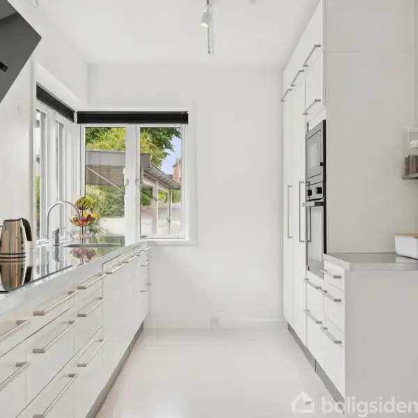 Et moderne køkken med hvide skabe, en indbygget ovn og en kogeplade. En elkedel står på bordpladen nær et vindue med udsigt til en baghave.