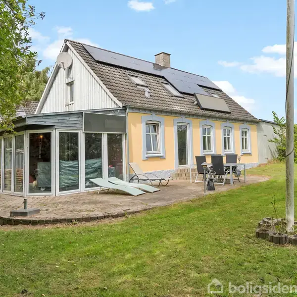 Et gult hus med solpaneler på taget står i en grøn have. En terrasse med liggestole og spisebord er ved siden af huset.