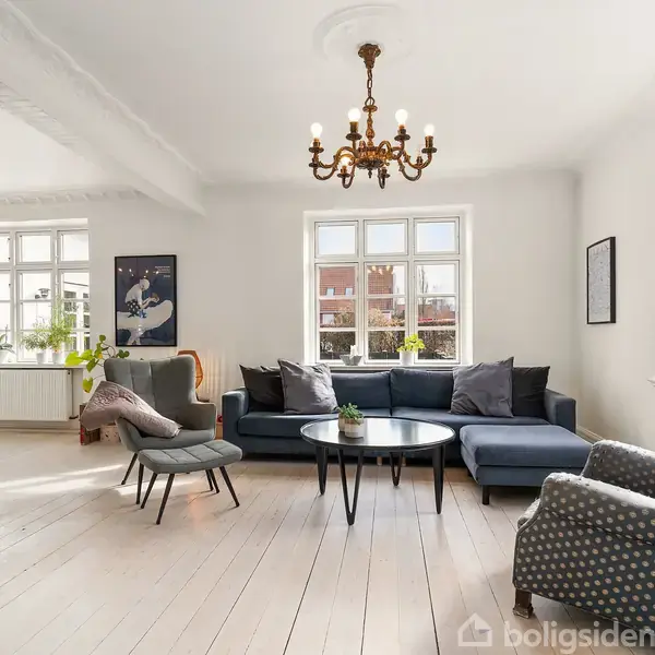 En blå sofa med puder står i en lys stue, flankeret af to mønstrede lænestole. En lysekrone hænger ned fra loftet, omgivet af vinduer og planter.