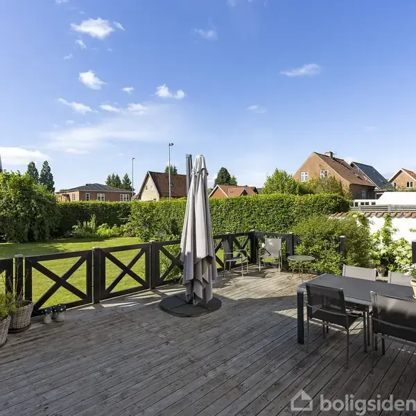 Terrasse med træbord og stole står i solen; en paraply er slået sammen ved siden af møblerne. Bagved ses en velplejet have og røde murstenshuse under en klar blå himmel.