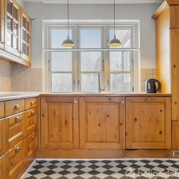 Køkken med træskabe og sort-hvid ternet gulv, oplyst af to loftslamper. Et vindue bagved tilbyder naturligt lys. En sort elkedel står på bordpladen.