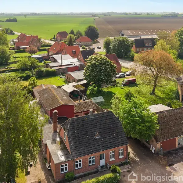 Murstenshus med sort tag står stille i en landsby omgivet af grønne træer og marker. Baggrunden viser flere huse og store, åbne marker under en blå himmel.