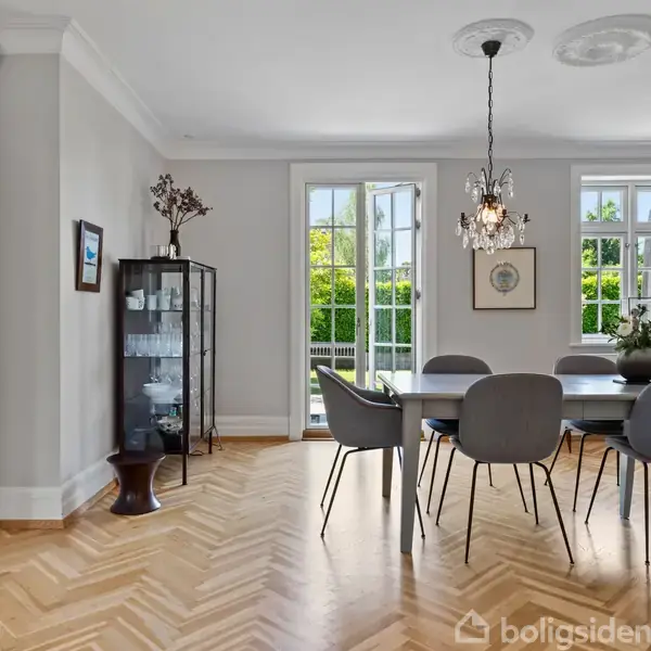 Et elegant spisebord med otte stole står under to lysekroner. Store vinduer med udsigt til en grøn have. Et vitrineskab med glas genstande i hjørnet. Ingen tekst.