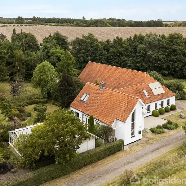 Hvidt hus med rødt tegltag ligger i et landligt område omgivet af træer og marker. Der er en grusvej, og en lille terrasse er placeret ved siden af huset.